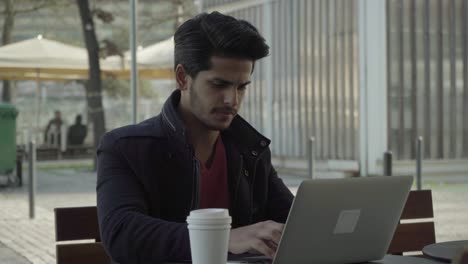 Serious-young-man-drinking-from-paper-cup-and-using-laptop